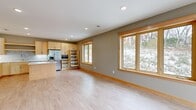 This well-appointed single-floor space showcases a clean, contemporary design with warm natural wood tones and light neutral wall colors throughout. The centerpiece is a spacious open-plan kitchen and living area, where light maple cabinetry, a tile mosaic backsplash, and stainless steel appliances — including a gas range and French door refrigerator — create a fully equipped culinary space. A central island with a white quartz countertop provides additional prep and gathering space. A built-in wine rack and pull-out pantry shelving add practical storage solutions. Recessed lighting illuminates the kitchen, complemented by light hardwood-style flooring that extends seamlessly throughout the space. Large wood-trimmed windows in the open living area frame views of a wooded, snow-dusted landscape, flooding the rooms with natural light. Additional rooms offer versatile layouts, each featuring the same warm wood trim, light flooring, ceiling fans, and windows with neighborhood and nature views. A dedicated laundry room houses a full-size washer and dryer with overhead shelving. The bathroom includes a clean white walk-in shower with brushed nickel fixtures. A stairwell with natural wood railings and windows provides additional brightness and connects to a lower entry. Overall, the space presents a cohesive, modern aesthetic with quality finishes throughout.