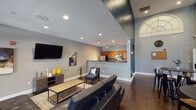 This well-appointed single-floor amenity space combines contemporary design with functional comfort throughout its open layout. The main lounge area is anchored by rich dark hardwood flooring, neutral gray walls, and recessed lighting that creates a warm, inviting atmosphere. A large wall-mounted flat-screen TV, a tufted leather sectional sofa, and a wooden coffee table make this an ideal gathering and relaxation area. French doors with transom windows open to an outdoor area, allowing natural light to pour in and providing views of the exterior grounds. A separate seating nook with a round bistro table and industrial-style stools sits beneath dramatically tall arched windows, adding architectural character and an abundance of daylight. The kitchen is fully equipped with warm wood cabinetry, dark granite countertops, stainless steel appliances including a refrigerator, microwave, and range, as well as a double basin sink and slate tile flooring. A peninsula with a dark granite overhang provides bar-style seating and seamlessly connects the kitchen to the living area. Track lighting throughout the kitchen ensures excellent task illumination. The overall design blends modern finishes with classic architectural details, resulting in a cohesive and welcoming communal environment.