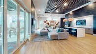 Located at 10210 Falcon Gate Loop in Riverview, FL, this single-story community clubhouse is thoughtfully designed to offer a dynamic mix of social, recreational, and productive spaces. The interior showcases a contemporary aesthetic throughout, highlighted by striking slatted wood ceiling treatments, recessed lighting, and wide-plank hardwood-style flooring that unifies the open layout.

The main lounge is a spacious, light-filled gathering area anchored by a large sectional sofa and centered around a dramatic floor-to-ceiling dark panel wall with a mounted flat-screen TV. Floor-to-ceiling glass doors open directly to an outdoor pool area, flooding the interior with natural light and seamlessly connecting indoor and outdoor living.

The game room is a standout feature, boasting a full-size billiards table, shuffleboard, ping-pong, and casual seating clusters beneath sculptural pendant lighting and a bold dark slatted ceiling. A built-in wet bar with sleek gray cabinetry, white quartz countertops, a stainless sink, and a large mounted display screen adds a refined entertainment element.

Private study rooms enclosed in glass partitions provide quiet, focused workspaces equipped with individual desks and chairs — ideal for remote work or personal productivity. The overall ambiance is upscale and inviting, balancing modern design with comfortable, community-centered functionality.