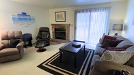 This single-floor space offers a warm and functional layout designed for comfortable everyday living. The living area serves as the focal point of the home, anchored by a classic brick fireplace with a wood mantle that adds a traditional, inviting character to the room. Natural light flows generously through a large sliding glass door dressed with vertical blinds, brightening the carpeted space and creating an open, airy feel. The living room is thoughtfully arranged with a plush sofa, a sleek black coffee table centered on a bold geometric area rug, and a leather recliner chair that adds both style and comfort. Wall art and decorative accents provide a personalized touch, while the neutral wall tones keep the atmosphere clean and understated. The overall ambiance is relaxed and homey, balancing practicality with comfort. The single-floor layout ensures easy accessibility and a seamless flow throughout the space, making it well-suited for a variety of lifestyles.