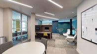 This single-floor amenity lounge is thoughtfully designed to support both productivity and relaxation. The space features a sophisticated palette of neutral tones accented by bold teal walls, patterned wallcovering, and pops of yellow in the furnishings, creating a dynamic yet cohesive atmosphere. Linear LED lighting runs across the ceiling throughout, providing bright, even illumination across all areas.

A built-in workstation area houses two individual computer stations set within a dark wood-finished unit, each equipped with a monitor and keyboard. Vibrant yellow chairs at the workstations add a lively contrast to the warm wood tones. Adjacent to this zone, a round white table with upholstered grey chairs offers a casual meeting or lounge area. Decorative wall art and sculpted accents contribute visual interest nearby.

A glass-walled meeting room provides a semi-private enclosed workspace, visible through full-height glazed partitions that maintain an open, connected feel. Toward one end of the space, a sleek white lounge chair with ottoman is positioned beside a glass-panel railing, overlooking a lower level finished in rich teal — adding architectural depth. Patterned tile flooring in varied grey tones anchors the entire space with texture and warmth.
