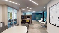 This single-floor amenity lounge is thoughtfully designed to support both productivity and relaxation. The space features a sophisticated palette of neutral tones accented by bold teal walls, patterned wallcovering, and pops of yellow in the furnishings, creating a dynamic yet cohesive atmosphere. Linear LED lighting runs across the ceiling throughout, providing bright, even illumination across all areas.

A built-in workstation area houses two individual computer stations set within a dark wood-finished unit, each equipped with a monitor and keyboard. Vibrant yellow chairs at the workstations add a lively contrast to the warm wood tones. Adjacent to this zone, a round white table with upholstered grey chairs offers a casual meeting or lounge area. Decorative wall art and sculpted accents contribute visual interest nearby.

A glass-walled meeting room provides a semi-private enclosed workspace, visible through full-height glazed partitions that maintain an open, connected feel. Toward one end of the space, a sleek white lounge chair with ottoman is positioned beside a glass-panel railing, overlooking a lower level finished in rich teal — adding architectural depth. Patterned tile flooring in varied grey tones anchors the entire space with texture and warmth.