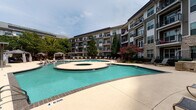 Located at 1300 Tribute Center Dr in Raleigh, NC, this outdoor amenity space delivers a resort-inspired poolside experience within a well-appointed residential community. The centerpiece is a generously sized freeform swimming pool with striking turquoise water, complemented by an integrated circular spa for relaxation. The pool's curvilinear design creates a dynamic, inviting atmosphere, and its shallow entry points and clearly marked depth areas ensure safety and accessibility for all users.

Surrounding the pool, a broad concrete sundeck offers ample space for lounging, furnished with rows of sleek reclining chairs that invite residents to soak up the sun. Shaded retreats are thoughtfully provided through a white pergola structure and multiple private cabanas, each curtained for privacy and outfitted with plush blue lounge chairs, side tables, and patio umbrellas — ideal for relaxing in comfort away from direct sunlight.

Lush landscaping featuring mature trees, flowering shrubs, and manicured greenery borders the pool area, adding vibrant color and natural beauty to the setting. Ornamental iron fencing defines the perimeter, lending a polished, secure feel to the space. The multi-story residential building wraps around the courtyard, offering balcony views overlooking this central outdoor retreat. The overall ambiance is one of a well-designed, resort-quality outdoor living space perfectly suited for leisure and socializing.