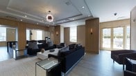 This refined single-floor space exudes contemporary elegance through a carefully curated blend of materials, lighting, and furnishings. The main lounge area commands immediate attention with its striking sputnik chandelier — a bold sculptural centerpiece suspended from a coffered ceiling accented by recessed lighting. Dark, plush sofas are arranged around a sleek marble coffee table, creating an inviting and sophisticated seating arrangement anchored by a neutral area rug.

Warm wood paneling lines the walls, lending a sense of richness and depth to the interior, while strategically placed wall sconces provide a soft ambient glow. Large windows allow natural light to filter into the space, offering views of the surrounding exterior and enhancing the overall brightness of the room. The flooring features wide-format stone or tile in a cool grey tone, providing a clean, modern contrast to the warmth of the wood elements.

Toward the rear, a mirrored wall expands the sense of space visually, reflecting the lounge and adding dimension. A separate area visible in the background suggests a dining or meeting zone, hinting at the multifunctional nature of the layout. The overall ambiance is polished and upscale, designed to impress while remaining functional and welcoming.