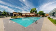 Located at 923 Maplewood Dr in Cedar Falls, Iowa, this outdoor recreational area serves as a vibrant community amenity designed for relaxation and leisure. The centerpiece of the space is a large, well-maintained swimming pool with crystal-clear blue water, lane markers, and ample deck space surrounding it on all sides. The expansive concrete pool deck is furnished with numerous lounge chairs and seating arrangements, including modern green-accented sofas and umbrellas that provide shaded areas for comfort on sunny days. The layout accommodates a generous number of residents, making it well-suited for both quiet relaxation and social gatherings. Enclosing the perimeter is a sturdy black iron fence that defines the space while maintaining an open, airy feel. Adjacent to the pool, thoughtfully designed landscaping features ornamental grasses, flowering ground cover, decorative boulders, and mature shrubs and trees that add natural beauty and a sense of privacy. A single-story clubhouse or utility building sits alongside the pool area, complementing the overall layout. The open sky above and the surrounding mature tree canopy contribute to a bright, inviting outdoor atmosphere. The space strikes a balance between functional amenity design and attractive, well-maintained grounds.