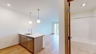 This thoughtfully designed 1-bedroom, 1-bathroom unit in Middleton, Wisconsin offers a functional and modern living experience across a single floor. The open-concept kitchen and living area forms the heart of the space, featuring warm light wood flooring that runs throughout. The kitchen is outfitted with stainless steel appliances — including a refrigerator, electric range, and over-the-range microwave — complemented by taupe cabinetry, light quartz countertops, and a peninsula with an undermount sink and pendant lighting overhead, creating a natural divide between the kitchen and living area. The living room is bright and airy, with clean white walls and sliding glass doors that open to a private balcony, offering pleasant outdoor views and an abundance of natural light. The bathroom features a full tub-shower combination with a curved curtain rod, light tile flooring, and a wood-toned vanity with gold hardware accents. A dedicated closet provides convenient storage. The unit's neutral color palette, consistent wood flooring, and modern fixtures create a cohesive, clean aesthetic throughout, making it a practical and comfortable residence in the Middleton area.