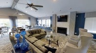 This single-story clubhouse offers a well-appointed communal space designed to accommodate both relaxed lounging and social gatherings. The main lounge area is anchored by a striking floor-to-ceiling fireplace clad in textured geometric tile, with a wall-mounted flat-screen TV positioned above it as a focal point. Vaulted ceilings fitted with a ceiling fan contribute to a sense of openness and airflow throughout the space.

The seating arrangement features plush, neutral-toned sofas and accent chairs arranged around a glass-top coffee table, all set atop a patterned area rug. Decorative blue glass accents add bold pops of color against the warm, earthy furnishings. Large windows dressed with layered curtains and blinds allow natural light to fill the interior while maintaining privacy.

Adjacent to the lounge, a casual dining area provides high-top tables and bar-style seating, ideal for informal conversations or light refreshments. The overall design blends contemporary and transitional styles, creating a welcoming and functional environment suited for community use.