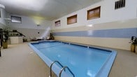 This indoor pool facility offers a welcoming aquatic environment designed for year-round use. The centerpiece is a generously sized rectangular pool with clear blue water, accessible via stainless steel entry rails and surrounded by a textured, slip-resistant deck. The pool's clean lines and well-maintained surface make it suitable for both recreational swimming and fitness laps.

The interior design features a combination of smooth upper walls in a neutral cream tone and a horizontal band of light blue ceramic tile along the mid-wall, adding a classic aquatic aesthetic. Above the tile line, a series of wood-slatted windows allow controlled natural light to filter into the space while maintaining privacy. Recessed and wall-mounted lighting supplements the ambient illumination throughout.

Along one side of the pool, a small seating area with chairs provides a comfortable space for relaxation or supervision. A potted plant adds a touch of greenery to the otherwise sleek environment. The drop ceiling contributes to a contained, cozy atmosphere while aiding in acoustics and climate control. Overall, the space is clean, functional, and thoughtfully designed for a comfortable swimming experience.
