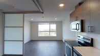 This modern single-floor space is designed with a clean, open-concept layout that maximizes both functionality and comfort. Dark hardwood flooring runs throughout, adding warmth and contrast against the crisp white walls and light-toned ceiling with recessed lighting. A large picture window serves as a focal point of the living area, flooding the space with natural light and offering sweeping views of the surrounding cityscape and treetops beyond. The open living area flows seamlessly into the kitchen, which features contemporary cabinetry in a rich wood finish, light quartz countertops, stainless steel appliances including a built-in microwave and electric range, and a sleek subway tile backsplash. A sliding door system with a modern track adds a thoughtful architectural detail, allowing for flexible division of space while maintaining an airy feel. Recessed lighting throughout ensures even illumination across the interior. The overall ambiance is fresh, minimalist, and urban — ideal for those who appreciate modern design paired with practical, well-appointed living spaces.
