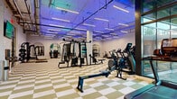 Located at 1788 Chain Bridge Rd in McLean, Virginia, this modern fitness center spans a generous single floor, offering a thoughtfully designed environment for a full range of workout activities. The main exercise floor is expansive and open, finished with a striking olive-and-white checkered tile floor that adds a bold graphic character to the space. Industrial-style exposed ceilings with linear LED lighting — accented by ambient blue uplighting — create an energetic yet polished atmosphere throughout. Large floor-to-ceiling windows along the perimeter flood the space with natural light and provide views to the exterior.

The main floor is comprehensively equipped with cardio machines including treadmills, ellipticals, rowing ergometers, and air bikes, alongside a wide array of strength equipment such as cable machines, power racks with TRX suspension systems, adjustable benches, and a full dumbbell rack. Arched wall niches with mirrors serve as decorative architectural accents that define different workout zones throughout the floor.

A separate studio room, enclosed by sleek black-framed glass partition walls, offers a serene space for yoga and stretching, featuring light hardwood flooring, high ceilings, and dramatic illuminated arched mirrors that reflect the room beautifully. A hydration station and display screen are also available for user convenience, rounding out this well-appointed facility.