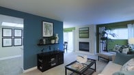 This single-floor space showcases a thoughtfully designed interior that balances bold color choices with a clean, contemporary aesthetic. The living area serves as the centerpiece, featuring a striking teal accent wall adorned with framed artwork and a sleek floating shelf displaying decorative objects and greenery. A dark-finished media console anchors the space, complementing the deep-toned furniture throughout.

A built-in fireplace with a modern surround adds a warm focal point to the room, positioned beneath an additional piece of wall art that echoes the cool, natural tones found throughout the space. The light-colored carpeting and bright white ceiling help to balance the deeper hues, creating an inviting and well-lit atmosphere.

Large sliding glass doors allow abundant natural light to fill the interior, framing an outdoor area and enhancing the sense of openness. Green Roman shades at the windows add a complementary pop of color to the teal and neutral palette. A glass-top coffee table with a dark frame sits at the center of the living area, adding a refined, airy touch. The overall ambiance is modern, cohesive, and comfortable, with carefully curated décor that gives the space a polished yet livable character.