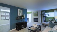 This single-floor space showcases a thoughtfully designed interior that balances bold color choices with a clean, contemporary aesthetic. The living area serves as the centerpiece, featuring a striking teal accent wall adorned with framed artwork and a sleek floating shelf displaying decorative objects and greenery. A dark-finished media console anchors the space, complementing the deep-toned furniture throughout.

A built-in fireplace with a modern surround adds a warm focal point to the room, positioned beneath an additional piece of wall art that echoes the cool, natural tones found throughout the space. The light-colored carpeting and bright white ceiling help to balance the deeper hues, creating an inviting and well-lit atmosphere.

Large sliding glass doors allow abundant natural light to fill the interior, framing an outdoor area and enhancing the sense of openness. Green Roman shades at the windows add a complementary pop of color to the teal and neutral palette. A glass-top coffee table with a dark frame sits at the center of the living area, adding a refined, airy touch. The overall ambiance is modern, cohesive, and comfortable, with carefully curated décor that gives the space a polished yet livable character.
