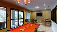 This single-floor recreational space is designed for entertainment and social gatherings. The centerpiece of the room is a full-sized pool table with a bold red felt surface, positioned beneath a cluster of warm pendant lights that create an inviting focal point. The lighting scheme combines recessed ceiling fixtures with the decorative pendants, ensuring the space is well-lit throughout.

Along one wall, a wall-mounted flat-screen television is framed by a built-in niche, accompanied by high-top tables and bar-height chairs, offering a comfortable area for viewing or casual dining. The neutral warm-toned walls complement the tile flooring, which features a subtle pattern that adds visual interest to the space.

Large glass doors and windows along one side of the room flood the interior with natural light while providing direct access to an outdoor patio area. Views through the glass reveal outdoor seating and surrounding greenery, creating a seamless connection between the indoor and outdoor environments. The overall ambiance is lively yet comfortable, making this space well-suited for social recreation.
