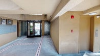 This single-story common area space serves as the communal hub of a residential complex, thoughtfully designed for both function and comfort. The covered entryway provides a generous shaded walkway with a textured stucco finish and concrete flooring accented by decorative tile inlays, offering residents a sheltered transition between the outdoors and the building interior. Cylindrical support columns frame the space, adding architectural structure while allowing open views of the surrounding greenery.

The secured entrance features glass-paned double doors flanked by glass block walls, allowing natural light to filter through while maintaining privacy and security. A fire alarm system is visibly mounted near the entry corridor, reflecting attention to safety standards throughout the building.

Adjacent to the entry is a dedicated mailbox area, housing a large bank of individual metal mailboxes for residents. Decorative framed mirrors and a wall-mounted lantern-style light fixture lend a welcoming, homey touch to this functional space.

Beyond the covered area, the courtyard opens up to reveal manicured hedges, mature trees, and well-kept lawn areas. Glimpses of a pool and additional residential buildings in the background suggest a broader community environment designed for outdoor enjoyment and comfortable communal living.
