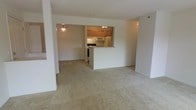 This single-floor residence offers a functional and inviting open-concept layout that seamlessly connects the living and kitchen areas. The spacious living area is finished with soft, neutral-toned wall-to-wall carpeting, creating a warm and comfortable atmosphere throughout the main living space.

The kitchen is positioned at the heart of the home, framed by warm honey-toned wood cabinetry that provides ample storage. A white peninsula counter separates the kitchen from the living area, offering a natural transition between spaces while also serving as a practical surface for dining or food preparation. The kitchen is equipped with a full suite of white appliances, including a refrigerator, microwave, and range, all set beneath under-cabinet lighting that brightens the workspace.

A pendant light fixture hangs above, adding a subtle decorative touch to the open layout. The clean, white walls throughout the space reflect light and contribute to an airy, uncluttered feel. The open sightlines from the living area into the kitchen and the adjacent space beyond enhance the sense of depth and openness, making this single-level home feel welcoming and easy to navigate.