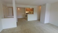 This single-floor residence offers a functional and inviting open-concept layout that seamlessly connects the living and kitchen areas. The spacious living area is finished with soft, neutral-toned wall-to-wall carpeting, creating a warm and comfortable atmosphere throughout the main living space.

The kitchen is positioned at the heart of the home, framed by warm honey-toned wood cabinetry that provides ample storage. A white peninsula counter separates the kitchen from the living area, offering a natural transition between spaces while also serving as a practical surface for dining or food preparation. The kitchen is equipped with a full suite of white appliances, including a refrigerator, microwave, and range, all set beneath under-cabinet lighting that brightens the workspace.

A pendant light fixture hangs above, adding a subtle decorative touch to the open layout. The clean, white walls throughout the space reflect light and contribute to an airy, uncluttered feel. The open sightlines from the living area into the kitchen and the adjacent space beyond enhance the sense of depth and openness, making this single-level home feel welcoming and easy to navigate.