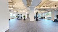 This expansive fitness and wellness facility offers a thoughtfully designed, single-floor layout that accommodates a wide range of exercise and wellness activities. The main exercise floor features durable rubber flooring and is generously sized, providing ample space for a full suite of cardio and strength training equipment, including stationary bikes, rowing machines, stair climbers, and cable machines. Structural columns punctuate the open floor plan, adding architectural character while preserving the spacious, airy feel of the room. Large windows line the perimeter, flooding the space with natural light and offering views of lush outdoor greenery.

A dedicated studio area with warm hardwood flooring provides an inviting environment for group fitness classes, yoga, barre, or stretching. This room features floor-to-ceiling glass partition walls with black framing, arched architectural details, wall-mounted mirrors, and a large window overlooking an outdoor garden. The exposed ceiling infrastructure throughout the facility, finished in white, adds an industrial-modern aesthetic while maintaining a bright and clean appearance. Utility rooms and storage areas are also accessible, ensuring full operational functionality. The overall ambiance is bright, clean, and energizing.