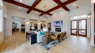 This well-appointed single-floor space functions as an inviting community lounge or clubhouse, thoughtfully designed to accommodate both relaxed socializing and productive work. The main living area immediately draws the eye upward to dramatic vaulted ceilings adorned with rich, dark wood cross-beams, complemented by a striking multi-bulb chandelier that anchors the room. Light hardwood flooring runs throughout, creating a warm and cohesive aesthetic.

The central seating area is anchored by a neutral sectional sofa accented with teal throw pillows, paired with leather-trimmed accent chairs and rustic wooden side tables. A wall-mounted flat-screen television sits above a classic fireplace, adding both function and a focal point to the lounge area. Expansive sets of French doors with dark wood framing line one wall, flooding the space with abundant natural light and providing access to the outdoors.

Adjacent to the main lounge is a well-equipped wet bar area featuring granite countertops, dark cabinetry, a beverage refrigerator, and a decorative patterned tile backsplash illuminated by accent lighting. Open floating shelves display curated décor alongside functional storage. A built-in counter workspace with a granite surface and Windsor-style wooden stools positioned beneath large windows offers an ideal spot for individual use. The overall ambiance is modern yet warm, blending upscale design elements with comfortable, community-oriented functionality.