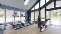 Located at 225 Echelon Rd in Voorhees, New Jersey, this fitness center is a well-appointed, single-floor facility designed to support a complete workout experience. The space immediately impresses with its dramatic vaulted ceiling, which rises to a striking peak and is accentuated by a skylight that floods the interior with natural light. Large floor-to-ceiling windows line the walls, offering expansive outdoor views and further enhancing the bright, open atmosphere of the room. The walls are finished in a deep navy blue, lending the space a sleek, modern aesthetic that pairs well with the commercial-grade equipment throughout. Multiple treadmills are positioned along the windowed perimeter, allowing users to exercise while enjoying the surrounding landscape. A cable strength machine and adjustable bench provide options for resistance training alongside the cardio equipment. Durable, neutral-toned carpeting covers the floor, providing comfort and stability underfoot. The combination of soaring architecture, generous natural light, and quality equipment makes this fitness center a functional and energizing environment for residents and users alike.