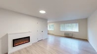 Located at 1000 W Ironwood Dr in Coeur d'Alene, Idaho, this thoughtfully updated single-floor unit offers a clean, modern aesthetic with a bright and welcoming atmosphere. The open living area is anchored by a sleek wall-mounted electric fireplace, providing both warmth and a contemporary focal point. Light luxury vinyl plank flooring flows seamlessly throughout the entire space, complementing the crisp white walls and creating a cohesive, airy feel. Natural light filters in through multiple windows fitted with horizontal blinds, offering views of the surrounding greenery. The living area is well-proportioned and connects to additional rooms that provide comfortable and versatile spaces, each featuring the same light flooring and neutral tones. A distinctive teal entry door adds a welcoming pop of color at the front entrance. The kitchen stands out with its two-tone cabinetry — pairing wood-grain upper cabinets with navy blue lower cabinets — set against bright white quartz countertops. Stainless steel appliances, including a refrigerator, range, and microwave, complete the kitchen's modern, functional design. Climate control is provided via wall-mounted HVAC units and baseboard heaters, ensuring year-round comfort throughout the space.