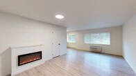 Located at 1000 W Ironwood Dr in Coeur d'Alene, Idaho, this thoughtfully updated single-floor unit offers a clean, modern aesthetic with a bright and welcoming atmosphere. The open living area is anchored by a sleek wall-mounted electric fireplace, providing both warmth and a contemporary focal point. Light luxury vinyl plank flooring flows seamlessly throughout the entire space, complementing the crisp white walls and creating a cohesive, airy feel. Natural light filters in through multiple windows fitted with horizontal blinds, offering views of the surrounding greenery. The living area is well-proportioned and connects to additional rooms that provide comfortable and versatile spaces, each featuring the same light flooring and neutral tones. A distinctive teal entry door adds a welcoming pop of color at the front entrance. The kitchen stands out with its two-tone cabinetry — pairing wood-grain upper cabinets with navy blue lower cabinets — set against bright white quartz countertops. Stainless steel appliances, including a refrigerator, range, and microwave, complete the kitchen's modern, functional design. Climate control is provided via wall-mounted HVAC units and baseboard heaters, ensuring year-round comfort throughout the space.