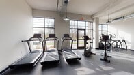 This fitness center is a comprehensive, single-floor facility designed to support a wide range of workout routines. The space is divided into distinct training zones, each thoughtfully outfitted to meet diverse exercise needs. The cardio area features multiple treadmills and upright stationary bikes arranged along floor-to-ceiling grid-pane windows, allowing ample natural light to pour in and offering views of the surrounding landscape — an energizing backdrop for any workout. A wall-mounted flat-screen television accompanies the treadmill section for added entertainment. The strength training zone is equipped with a variety of selectorized cable machines, multi-functional weight stations, adjustable benches, and a full dumbbell rack, providing options for both targeted muscle training and compound movements. A dedicated functional training area features hardwood-style flooring and is stocked with stability balls, resistance bands, medicine balls, a plyo box, and a rowing machine — ideal for dynamic and mobility-focused workouts. Throughout the facility, exposed ductwork and pendant industrial-style lighting contribute to a modern, warehouse-inspired aesthetic. Durable rubber flooring covers the primary workout areas, ensuring safety and comfort. The space is bright, open, and efficiently organized to maximize usability across all fitness levels.