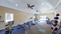 This fitness center is a thoughtfully designed, single-floor facility offering a comprehensive range of exercise equipment to support a full-body workout routine. The space features warm, neutral-toned walls accented by crisp crown molding, contributing to a clean and welcoming atmosphere throughout.

The cardio zone is well-appointed with multiple treadmills and elliptical trainers arranged to maximize floor space, while stationary bikes provide additional low-impact options. Natural light flows in generously through several windows positioned along the walls, creating a bright and energizing environment. Multiple ceiling fans with recessed lighting ensure comfortable air circulation during workouts.

A dedicated strength training area houses a full rack of dumbbells in varying weights alongside medicine balls and a multi-use adjustable bench, catering to both beginners and experienced athletes. The durable dark-tiled flooring with accent mat sections provides stability and defines workout zones throughout the room.

Wall-mounted flat-screen televisions are positioned for easy viewing from the cardio equipment, adding entertainment to the exercise experience. The space is finished with safety features including fire extinguishers and clearly marked exits, ensuring a safe and functional environment. Entry doors provide convenient access, completing this well-rounded fitness facility.