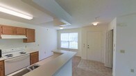 This single-floor space offers a straightforward, functional layout designed for comfortable everyday living. The living area is finished with neutral-toned wall-to-wall carpeting that extends through the main rooms, creating a cohesive and understated aesthetic. Crisp white walls and flush-mounted ceiling lights keep the space feeling clean and bright throughout.

An open passageway connects the living area to a secondary room, both benefiting from natural light streaming through large windows. The windows provide a generous source of daylight, contributing to the airy feel of the interior.

The kitchen is positioned adjacent to the living and dining area and features a peninsula-style breakfast bar, offering both prep space and casual seating potential. Warm wood-toned cabinetry lines the kitchen walls, complemented by a built-in sink. The kitchen and entry areas are finished with tile flooring, offering a practical contrast to the carpeted living spaces.

The front entry is defined by a paneled white door and tile flooring, creating a clear transition into the home. A textured ceiling adds subtle visual interest to the kitchen and dining area. Overall, the space presents a clean, move-in ready environment with a simple, functional design.