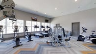 This fitness center offers a well-rounded workout environment designed for both cardio and strength training. The space is finished with a neutral gray and white color palette, contributing to a clean, modern aesthetic. A patterned low-pile carpet in varying shades of gray and tan covers the floor, adding visual interest while providing a practical surface underfoot.

Large windows line one wall, flooding the interior with natural light and offering views of surrounding greenery, creating an open and inviting atmosphere. Recessed ceiling lighting supplements natural light for evening use. Two wall-mounted flat-screen televisions are positioned for convenient viewing during workouts.

The cardio section is well-stocked, featuring multiple treadmills and elliptical machines aligned along the windowed wall. Strength training equipment, including a multi-station cable machine and a leg press or seated resistance machine, is positioned toward the center and rear of the room. A recumbent bike rounds out the cardiovascular offerings. A small counter area near the rear provides a functional utility space. The overall layout is thoughtfully organized, ensuring adequate spacing between equipment for comfortable, safe use.