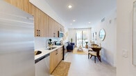 Located at 208 N Federal Hwy in Pompano Beach, FL, this thoughtfully designed one-bedroom, one-bathroom residence offers a bright and cohesive coastal aesthetic throughout. The open-concept kitchen and living area form the heart of the home, where warm wood-tone cabinetry with matte black hardware pairs seamlessly with white quartz countertops and a classic subway tile backsplash. Stainless steel appliances, including a full-size refrigerator, dishwasher, and range, complete the well-appointed kitchen. Light porcelain tile flooring flows throughout the main living area, reflecting the abundant natural light that pours in through a sliding glass door. This door opens to an outdoor balcony with views of palm trees, reinforcing the Florida coastal atmosphere. The living and dining area is finished with crisp white walls and recessed lighting, creating an airy, welcoming ambiance. The bedroom is a tranquil retreat, featuring a ceiling fan, a window with lush outdoor greenery views, and soft neutral tones. A dedicated closet provides convenient storage. The bathroom is elegantly finished with floor-to-ceiling marble-look tile in a frameless glass walk-in shower, delivering a clean, upscale feel. A hallway connects all areas of the residence efficiently.