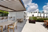 Located at 20775 NW 17th Ave in Miami Gardens, Florida, this rooftop amenity deck offers a well-appointed open-air experience designed for both relaxation and social gathering. The space is defined by a clean, modern aesthetic with white architectural walls, light-toned paver flooring, and a striking louvered pergola that provides partial shade over an outdoor kitchen and dining area. The pergola-covered zone features a built-in grill station alongside multiple seating arrangements, including wood-and-canvas chairs and round dining tables suited for group gatherings. Adjacent to this covered section, the open-air deck extends across a generous expanse, lined with large raised planters filled with vibrant tropical and ornamental greenery — including agave, succulents, and colorful foliage — that add a lush, garden-like character to the elevated setting. A dedicated section of the rooftop features a well-maintained turf lawn area, offering a versatile green space ideal for casual recreation or relaxation. Lounge seating with cushioned benches is thoughtfully positioned throughout to take advantage of the open sky and sweeping views of the surrounding Miami Gardens landscape. The entire deck is enclosed by sleek metal railings that maintain unobstructed sightlines to the horizon, creating a bright and airy ambiance under South Florida's expansive sky.