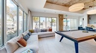 Located at 265 Avenue E SW in Winter Haven, Florida, this thoughtfully designed single-floor amenity space offers a generous, open layout that seamlessly blends function with modern coastal aesthetics. Light-toned large-format tile flooring flows throughout, unifying the various areas under soaring ceilings that feature an eye-catching exposed concrete finish accented by bold drum pendant lighting.

The expansive main living area is anchored by a stylish seating arrangement — a neutral sectional sofa and leather accent chairs paired with a coastal-inspired large-format wall art piece, creating a warm and inviting gathering environment. Adjacent to this, a second lounge area provides flexible seating with views to an outdoor terrace accessible through full-height sliding glass doors, flooding the space with natural light and views of mature palm and oak trees.

The well-appointed kitchen features a long quartz waterfall island with bar seating, textured grey cabinetry, a white tile backsplash, and sleek pendant lighting. A dedicated beverage and coffee station adds everyday convenience. The dining area incorporates a walnut-toned table beneath an exposed concrete ceiling, accompanied by a wall-mounted display screen — ideal for gatherings or presentations. Private glass-fronted office rooms and a formal conference room with additional AV capabilities round out this versatile, professionally finished space.