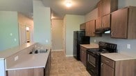 This single-floor space is designed with practicality and comfort in mind. The kitchen serves as a central highlight, featuring a classic galley layout that maximizes efficiency. Rich, dark wood cabinetry lines both sides of the kitchen, offering generous storage through upper and lower cabinet configurations. The kitchen is equipped with black appliances, including a full-size refrigerator, a gas range with oven, and a double-basin sink with updated fixtures set into a light-toned countertop. The earthy, stone-patterned tile flooring adds a grounded, durable quality to the space and flows seamlessly throughout the kitchen and into the adjoining hallway. Soft sage-green walls contribute a calm, neutral ambiance that pairs well with the warm wood tones of the cabinetry. Overhead lighting keeps the kitchen well-lit, while a hallway visible from the kitchen leads further into the unit, hinting at additional living spaces beyond. White baseboards and trim provide clean, crisp contrast against the walls, lending the interior a tidy, well-maintained appearance. Overall, the space offers a straightforward, functional layout with cohesive design choices throughout.