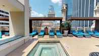 Located in the heart of downtown Dallas at 1509 Main St, this rooftop amenity deck offers an impressive outdoor retreat elevated above the city streetscape. The centerpiece of the space is a generously sized swimming pool with clear turquoise water, flanked by a wide wood-plank deck that provides ample room for relaxation and movement. Adjacent to the pool, a spa hot tub offers a more intimate soaking experience, also set within the same deck surface for a cohesive layout.

The lounge area is well-appointed with multiple cushioned chaise lounges finished in vibrant blue, arranged beneath a sturdy pergola structure that provides partial shade during sunny Texas days. Additional seating, including patio chairs and tables, allows for casual gathering and socializing. Ornate architectural columns with decorative figurines accent the perimeter of the deck, reflecting the classical design heritage of the surrounding historic Dallas architecture visible from the rooftop.

The space is framed on all sides by panoramic views of the downtown Dallas skyline, with prominent glass towers rising in the background, offering a dramatic urban backdrop. Large glass doors provide interior access to enclosed amenity spaces. Potted trees and greenery add natural warmth to the otherwise sleek and modern outdoor environment.