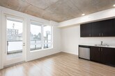 This contemporary single-floor unit showcases a thoughtfully designed open-concept layout that seamlessly connects the main living areas. Light hardwood flooring runs throughout the space, creating a warm and cohesive aesthetic that complements the clean white walls.

The kitchen is a standout feature, outfitted with rich dark cabinetry — available in both deep espresso brown and navy tones — paired with white countertops and a full suite of stainless steel appliances, including an electric range, built-in microwave, dishwasher, and refrigerator. The kitchen opens directly into the living area, which benefits from natural light streaming in through a window with views of the surrounding neighborhood.

A hallway lined with sliding barn doors on a sleek metal track leads to the private areas of the unit, adding a modern industrial touch. The exposed concrete ceiling in portions of the space further enhances the contemporary loft-like character, complemented by recessed lighting.

The bedroom is accessed via a sliding barn door and features durable hardwood-style flooring and ample wall space. The bathroom is clean and functional, featuring a large vanity with dark cabinetry, white countertop, a wide mirror with overhead lighting, and light hardwood flooring. The overall ambiance is modern, minimal, and move-in ready.