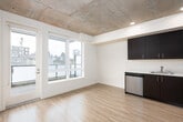 This contemporary single-floor unit showcases a thoughtfully designed open-concept layout that seamlessly connects the main living areas. Light hardwood flooring runs throughout the space, creating a warm and cohesive aesthetic that complements the clean white walls.

The kitchen is a standout feature, outfitted with rich dark cabinetry — available in both deep espresso brown and navy tones — paired with white countertops and a full suite of stainless steel appliances, including an electric range, built-in microwave, dishwasher, and refrigerator. The kitchen opens directly into the living area, which benefits from natural light streaming in through a window with views of the surrounding neighborhood.

A hallway lined with sliding barn doors on a sleek metal track leads to the private areas of the unit, adding a modern industrial touch. The exposed concrete ceiling in portions of the space further enhances the contemporary loft-like character, complemented by recessed lighting.

The bedroom is accessed via a sliding barn door and features durable hardwood-style flooring and ample wall space. The bathroom is clean and functional, featuring a large vanity with dark cabinetry, white countertop, a wide mirror with overhead lighting, and light hardwood flooring. The overall ambiance is modern, minimal, and move-in ready.