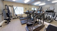 This fitness center offers a focused and functional environment designed to support a well-rounded workout routine. The space is finished with neutral, light-toned walls and durable flooring, creating a clean and practical atmosphere conducive to exercise. Overhead fluorescent lighting ensures the room is well-lit throughout the day, complemented by natural light streaming in through white-trimmed windows that overlook a lush, green outdoor setting.

The cardio section is a highlight of the space, featuring multiple Landice treadmills and an elliptical trainer — brands recognized for their quality and durability. A multi-station cable and weight machine is positioned along one wall, providing options for strength and resistance training. Full-length mirrors line one wall, visually expanding the space and allowing users to monitor their form during workouts.

The layout is thoughtfully organized to maximize usability without overcrowding, allowing for comfortable movement between equipment. Framed artwork on the walls adds a subtle decorative touch, keeping the environment welcoming rather than purely utilitarian. Overall, this fitness center delivers a well-appointed and inviting space for residents or guests seeking convenient access to quality exercise facilities.
