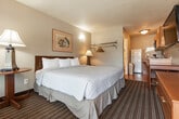 This single-floor studio guest room is efficiently designed to accommodate both short-term and extended stays. The main living area is centered around a queen-sized bed dressed in neutral-toned bedding, flanked by a bedside lamp that provides warm ambient lighting. Framed wall art and a wall-mounted clothing rack with hangers add functional and decorative touches. Patterned carpeting covers the sleeping area, while wood-look flooring transitions into the kitchenette zone.

The kitchenette is well-equipped with warm oak cabinetry, a countertop range, microwave, full-size refrigerator, stainless steel sink, and a coffee maker — offering convenient in-room meal preparation. A small dining table and chair are positioned nearby. A wall-mounted flat-screen television and a multi-drawer wooden dresser are situated across from the bed, enhancing the room's utility.

A large window near the entry door allows natural light to fill the space, fitted with striped curtains for privacy. The bathroom features a white vanity with a single sink, a framed mirror, and a full tub and shower combination with tan tile surround. Tile flooring and clean white walls complete the bathroom's practical, no-frills design.