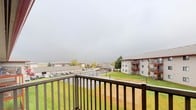 This large balcony is a standout outdoor feature that provides a generous amount of usable exterior space. Situated on an upper floor, it offers a broad, elevated vantage point overlooking the surrounding community. The balcony is enclosed by a sturdy dark-railed railing system, providing both safety and an unobstructed sightline across the landscape below.

The views from the balcony extend across well-maintained green lawns, a parking area, and on-site tennis courts, with neighboring residential buildings and mature trees visible in the distance. The wide, open sky above further enhances the sense of openness and airiness that this outdoor space provides.

The balcony deck features wood-toned flooring, lending a warm, natural aesthetic to the space. Its generous dimensions make it well-suited for outdoor furniture, allowing residents to create a comfortable seating or dining area for enjoying the outdoors. Whether used as a quiet retreat or a space for casual outdoor entertaining, this balcony offers a meaningful connection to the outdoors and the surrounding neighborhood environment.