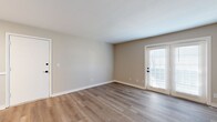 This thoughtfully designed single-story unit offers a comfortable and functional layout across one floor. The combined kitchen and living area forms the heart of the home, with the living space featuring warm wood-look luxury vinyl plank flooring, neutral beige walls, and a set of French doors that invite natural light and provide direct outdoor access. The kitchen is a standout, boasting crisp white shaker-style cabinetry with sleek matte black hardware, light quartz countertops, a stainless steel sink with a matte black gooseneck faucet, and a full suite of stainless steel appliances. Ample upper and lower cabinet storage ensures excellent functionality. A hallway connects the main living areas to the private bedroom and bathroom. The generously sized bedroom is carpeted for comfort and includes a window with blinds that allows for natural light. A dedicated closet provides additional storage. The full bathroom features a striking marble-look tub surround, a white vanity with quartz countertop and matte black fixtures, and wood-look flooring that ties the space together cohesively. Clean lines, consistent neutral tones, and quality finishes throughout give this unit a fresh, move-in-ready appeal.