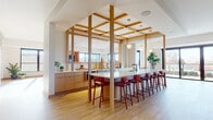 This thoughtfully designed single-floor space blends modern architecture with warm, inviting interiors across a variety of distinct functional zones. Light hardwood flooring flows continuously throughout, creating visual cohesion between areas.

The centerpiece is a striking open kitchen anchored by a long white-topped island lined with bold red barstools. Vertical wood columns support an eye-catching overhead grid of natural wood beams, framing the kitchen zone while maintaining an open connection to surrounding areas. Flat-front warm wood cabinetry lines the walls alongside integrated appliances, and globe pendant lights add a refined touch.

Adjacent lounge areas offer varied seating experiences — from a sunlit window-side platform with built-in upholstered benches overlooking an outdoor terrace, to a relaxed media lounge furnished with deep burgundy sectional sofas and dual wall-mounted televisions. A casual dining nook features patterned accent walls, banquette seating, and woven pendant lighting for an intimate atmosphere.

A game room with a shuffleboard table and bold chevron-patterned feature wall adds a social dimension, while glass-walled meeting rooms provide a transparent, semi-private workspace. Throughout, greenery, curated artwork, and a consistent warm palette of terracotta, burgundy, and natural wood create a cohesive and welcoming ambiance.