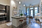 Located at 2245 Interstate North Pkwy SE in Atlanta, Georgia, this single-floor residence offers a well-designed layout that balances style and functionality. The open-concept kitchen serves as a central highlight, showcasing dark shaker-style cabinetry, granite countertops, a full suite of stainless steel appliances, and a subway tile backsplash. A large central island with pendant lighting and bar seating creates an ideal space for casual dining and entertaining. The soaring ceilings in the main living area amplify the sense of space, while wood-style flooring runs throughout the common areas, adding warmth and continuity. The living area is bright and inviting, with large windows that frame lush outdoor greenery and allow abundant natural light to fill the room. The bedrooms are comfortably scaled, each featuring plush carpeting, large windows with tree-lined views, and ample wall space. The primary suite connects directly to a well-appointed en-suite bathroom, which features a granite-topped double vanity with dark cabinetry, wood-look tile flooring, a glass-enclosed shower, and direct access to a generously sized walk-in closet outfitted with wire shelving systems. The overall aesthetic is clean and contemporary throughout.