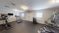 Located at 1601 NW 13th St in Boca Raton, Florida, this dedicated fitness center offers a well-equipped, single-room workout space designed to support a comprehensive exercise routine. The room features durable rubber flooring throughout, providing both safety and comfort underfoot, while crown molding and soft gray walls lend a clean, polished aesthetic to the interior.

Overhead flush-mount lighting illuminates the space evenly, complemented by natural light filtering in through multiple windows fitted with white blinds. A wall-mounted flat-screen television adds a convenient entertainment option during workouts, and a wall mirror enhances the sense of openness.

The fitness center is stocked with a well-rounded selection of equipment. Cardio options include a treadmill and an elliptical trainer, while strength training is supported by a multi-station cable machine, a dedicated weight bench, and a full rack of dumbbells ranging across various weights. Stability and functional training tools such as medicine balls, a BOSU balance trainer, and exercise balls round out the offerings. A motivational "fitness" word art decal adorns one wall, reinforcing the room's purpose.

Access is provided through a set of French doors as well as a solid double-door entry, both secured and clearly marked with exit signage. This turnkey fitness facility is ideally suited for residents seeking convenient, on-site access to a fully functional gym.