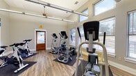 This fitness center is a purpose-built exercise space designed to support a full range of cardio and functional training workouts. The room features high ceilings with elevated clerestory windows that allow abundant natural light to fill the space, complemented by large lower windows with blinds that offer views of the surrounding outdoor greenery. The flooring is durable wood-look planking, with protective rubber mats positioned beneath each piece of equipment.

The facility is outfitted with a varied selection of cardio machines, including multiple elliptical trainers, spinning bikes, and a low-impact cross trainer, providing options for users of all fitness levels. A wall-mounted suspension training system with resistance straps adds a functional training component to the space. Overhead, industrial-style heating units and a ceiling fan ensure year-round comfort during workouts.

A striking motivational typography mural dominates one wall, featuring bold blue and white lettering with phrases such as "Keep Fit," "Train Stronger," and "Live Strong," creating an energizing and focused atmosphere. A warm-toned wooden entry door serves as the main access point. The overall design is clean, open, and functional, striking a balance between practicality and an inviting workout environment.