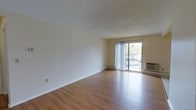 This single-floor 1-bedroom, 1-bathroom residence offers a clean, functional layout designed for comfortable everyday living. The living area is spacious and open, featuring warm hardwood-style flooring that runs throughout and crisp, neutral-toned walls that create a bright and inviting atmosphere. A large sliding glass door floods the space with natural light and provides direct access to a private balcony, offering views of the surrounding greenery and streetscape beyond. A baseboard heating unit and a wall-mounted climate control unit ensure year-round comfort. The overall design is simple and understated, with smooth ceilings and clean lines that lend the space a versatile, move-in-ready character. The open feel of the living area makes it easy to arrange and personalize. With its natural light, outdoor access, and practical layout, this residence is well-suited for those seeking a straightforward and comfortable living space.