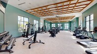 This expansive fitness center offers a generously sized, single-floor workout facility designed to accommodate a full range of exercise needs. The space immediately impresses with its distinctive architectural ceiling feature — a bold wood lattice grid suspended overhead, lending warmth and visual depth to the interior. Soaring ceiling heights enhance the sense of openness throughout.

The walls are finished in a rich teal hue accented by crisp white upper sections, creating an energizing yet refined atmosphere. Large, floor-to-ceiling windows line multiple walls, flooding the space with abundant natural light and offering views of the surrounding outdoor landscape, including palm trees and greenery.

The facility is comprehensively equipped with a wide selection of strength training machines, including cable systems, multi-function press and fly machines, and a functional training rig with TRX suspension equipment. The cardio zone features treadmills, stationary bikes, rowing machines, and stair climbers, all with modern digital displays. Free weight benches and additional resistance stations round out the strength offerings.

The flooring consists of durable, light-toned tile throughout, providing a clean and practical surface for all workout activities. Ceiling fans and a mounted flat-screen television add to the comfort and convenience of the space. Large mirrors in select zones support proper form during exercise. The overall design balances aesthetic appeal with practical, high-performance fitness functionality.