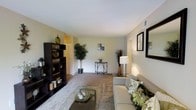This single-floor residence offers a comfortable and functional layout designed for everyday living. The living room serves as the heart of the space, showcasing a warm, neutral color palette with soft beige walls that create a calm and inviting atmosphere. Plush wall-to-wall carpeting adds a sense of coziness underfoot, while the open layout allows for generous furniture arrangement and easy movement throughout the room. A dark-toned bookcase along one wall provides both storage and a stylish focal point, complementing the understated elegance of the space. Decorative indoor plants introduce natural elements, adding life and texture to the interior. A central console table with accent décor draws the eye toward the far wall, while framed artwork displayed on the right wall adds a refined, finished touch. Recessed wall vents and smooth ceilings contribute to the clean architectural lines of the space. Natural and artificial lighting work in harmony, with a classic table lamp providing warm, focused illumination. A hallway visible in the background hints at additional rooms, suggesting a thoughtfully organized floor plan. Overall, this space combines practicality with a welcoming aesthetic, making it an ideal environment for comfortable, day-to-day living.