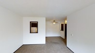 This single-floor residential space offers a straightforward and functional layout designed for comfortable everyday living. The main living area is finished with wall-to-wall carpeting that provides warmth underfoot, while white walls keep the space feeling bright and clean. A ceiling fan is centrally positioned overhead, offering both light and air circulation throughout the room.

A defining architectural detail is the pass-through window cut into the interior wall, which connects the living area directly to the kitchen. This feature enhances the open feel of the space while maintaining a visual separation between the two areas. Through the pass-through, the kitchen is visible and includes dark-toned cabinetry and a white refrigerator, suggesting a compact but well-organized culinary space.

A hallway visible toward the back of the living area leads further into the unit, hinting at additional rooms beyond. Dark wood trim accents frame the doorways and pass-through window, providing contrast against the white walls and adding a touch of definition to the interior. The textured ceiling adds subtle visual character to the overhead plane. Overall, the space presents a clean, no-frills environment with practical design choices suited for daily residential use.