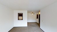 This single-floor residential space offers a straightforward and functional layout designed for comfortable everyday living. The main living area is finished with wall-to-wall carpeting that provides warmth underfoot, while white walls keep the space feeling bright and clean. A ceiling fan is centrally positioned overhead, offering both light and air circulation throughout the room.

A defining architectural detail is the pass-through window cut into the interior wall, which connects the living area directly to the kitchen. This feature enhances the open feel of the space while maintaining a visual separation between the two areas. Through the pass-through, the kitchen is visible and includes dark-toned cabinetry and a white refrigerator, suggesting a compact but well-organized culinary space.

A hallway visible toward the back of the living area leads further into the unit, hinting at additional rooms beyond. Dark wood trim accents frame the doorways and pass-through window, providing contrast against the white walls and adding a touch of definition to the interior. The textured ceiling adds subtle visual character to the overhead plane. Overall, the space presents a clean, no-frills environment with practical design choices suited for daily residential use.