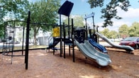 Located at 3001 Reflections Way in Virginia Beach, Virginia, this community space combines an inviting outdoor playground with a well-appointed interior entry corridor. The outdoor play area is a standout feature, showcasing a robust multi-level play structure finished in a sleek black frame. It includes multiple slides, a climbing tower, interactive activity panels, and a freestanding circular climbing net — all set over a wood chip safety surface. Mature trees provide generous shade across the play area, creating a comfortable and sheltered environment for outdoor activity beneath open Virginia skies. The interior entry corridor offers a clean, welcoming transition into the building. Stone-look ceramic tile flooring in warm earth tones runs throughout the hallway, complemented by crisp white paneled doors and neutral warm-toned walls. Nautical-themed framed artwork depicting serene coastal and sailing scenes lines the walls, nodding to the community's Virginia Beach surroundings. Glass-panel entry doors allow natural light to filter into the space, enhancing its airy feel. Safety features including a clearly marked fire alarm pull station are also present. Together, the outdoor and indoor spaces reflect a community designed with both functionality and coastal character in mind.